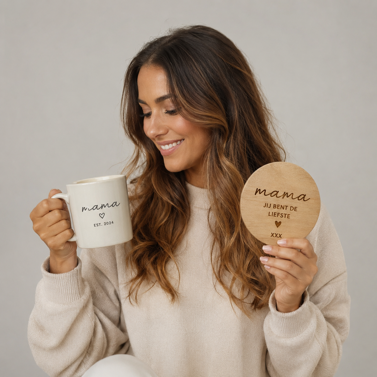 Vrolijke vrouw met gepersonaliseerde mok en gegraveerd houten cadeau voor mama, gefotografeerd in een luxe studio met neutrale beige en crèmekleuren. Perfect voorbeeld van een origineel gepersonaliseerd cadeau voor Moederdag, verjaardag of speciale gelegenheid.