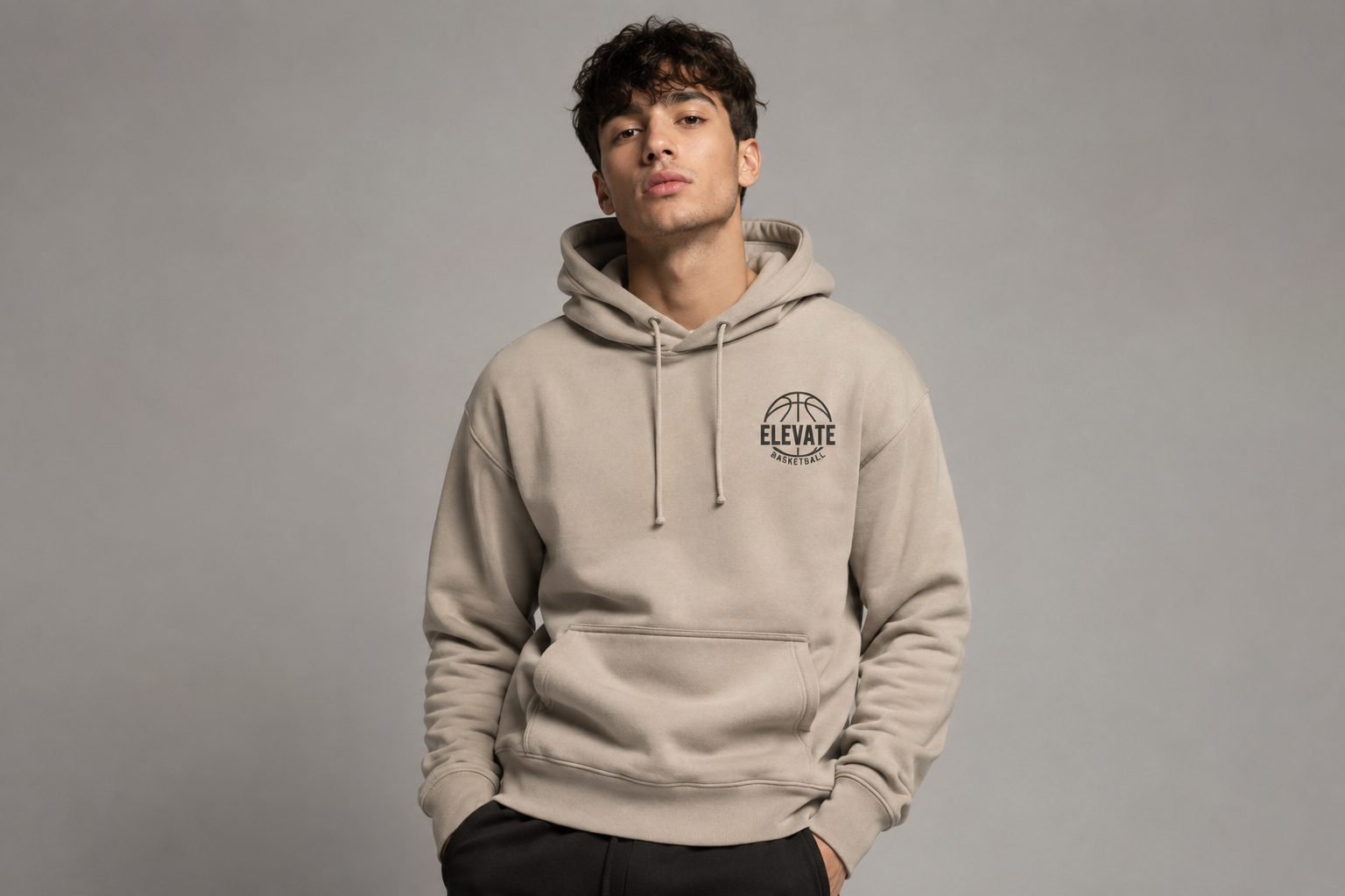 Jongvolwassene draagt een bedrukte beige team hoodie met subtiel logo op een lichtgrijze studioachtergrond. Professionele bedrijfskleding en gepersonaliseerde hoodies voor teams, verenigingen en bedrijven, gefotografeerd in een moderne premium e-commerce stijl.